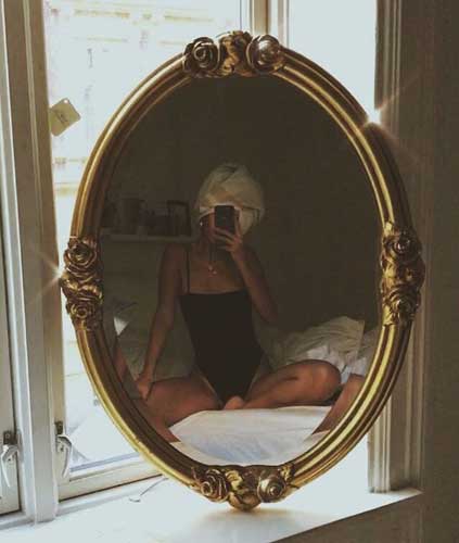 espejo antiguo que refleja la imagen de una mujer sentada en una cama con un body negro y una toalla blanca en su cabeza